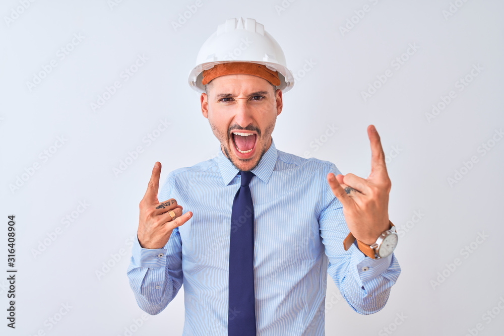Young business man wearing contractor safety helmet over isolated background shouting with crazy expression doing rock symbol with hands up. Music star. Heavy concept. Young business man wearing contractor safety helmet over isolated background shouting with crazy expression doing rock symbol with hands up. Music star. Heavy concept.