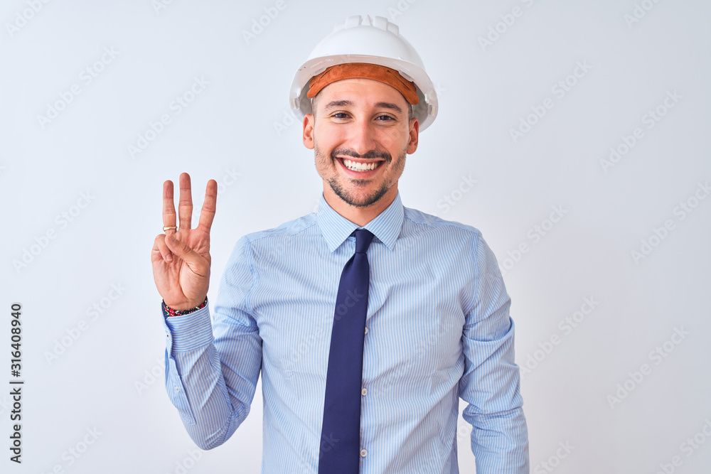 Young business man wearing contractor safety helmet over isolated background showing and pointing up with fingers number three while smiling confident and happy.