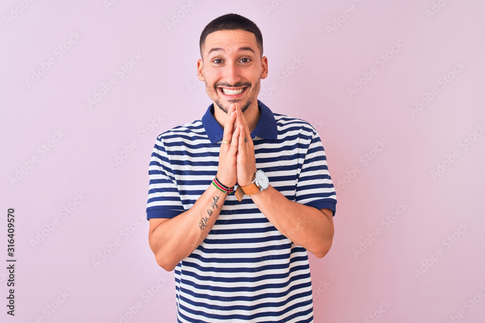 Young handsome man wearing nautical striped t-shirt over pink isolated background praying with hands together asking for forgiveness smiling confident. Young handsome man wearing nautical striped t-shirt over pink isolated background praying with hands together asking for forgiveness smiling confident.