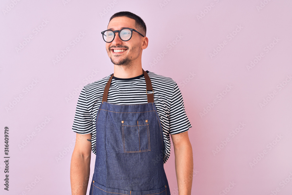 Young handsome man wearing employee apron over isolated background looking away to side with smile on face, natural expression. Laughing confident. Young handsome man wearing employee apron over isolated background looking away to side with smile on face, natural expression. Laughing confident.