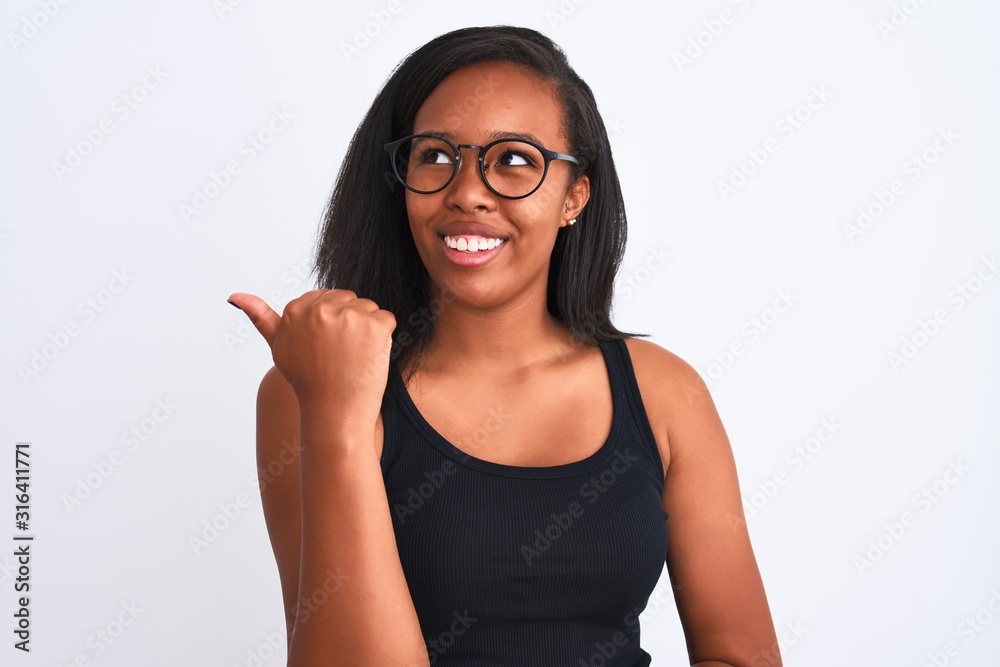 Beautiful young african american woman wearing glasses over isolated background smiling with happy face looking and pointing to the side with thumb up. Beautiful young african american woman wearing glasses over isolated background smiling with happy face looking and pointing to the side with thumb up.