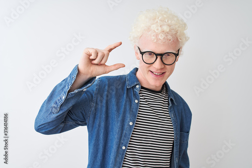 Young albino blond man wearing denim shirt and glasses over isolated white background smiling and confident gesturing with hand doing small size sign with fingers looking and the camera