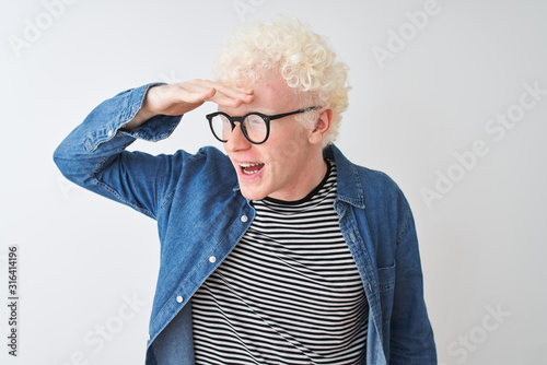 Young albino blond man wearing denim shirt and glasses over isolated white background very happy and smiling looking far away with hand over head. Searching concept.