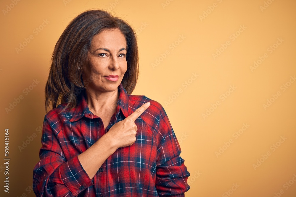 Middle age beautiful woman wearing casual shirt standing over isolated yellow background Pointing with hand finger to the side showing advertisement, serious and calm face Middle age beautiful woman wearing casual shirt standing over isolated yellow background Pointing with hand finger to the side showing advertisement, serious and calm face
