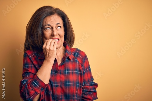 Middle age beautiful woman wearing casual shirt standing over isolated yellow background looking stressed and nervous with hands on mouth biting nails. Anxiety problem.