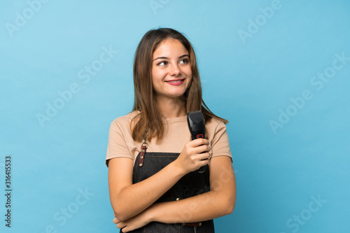 Young brunette girl over isolated blue background with hairdresser or barber dress and holding hair cutting machine