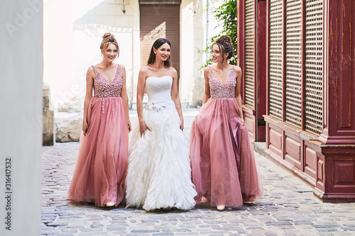 Beautiful bride and bridesmaids walking on the cobblestone street. Maids wear gorgeous elegant stylish red pink violet floor length v neck chiffon gown dress decorated with sequins sparkles and