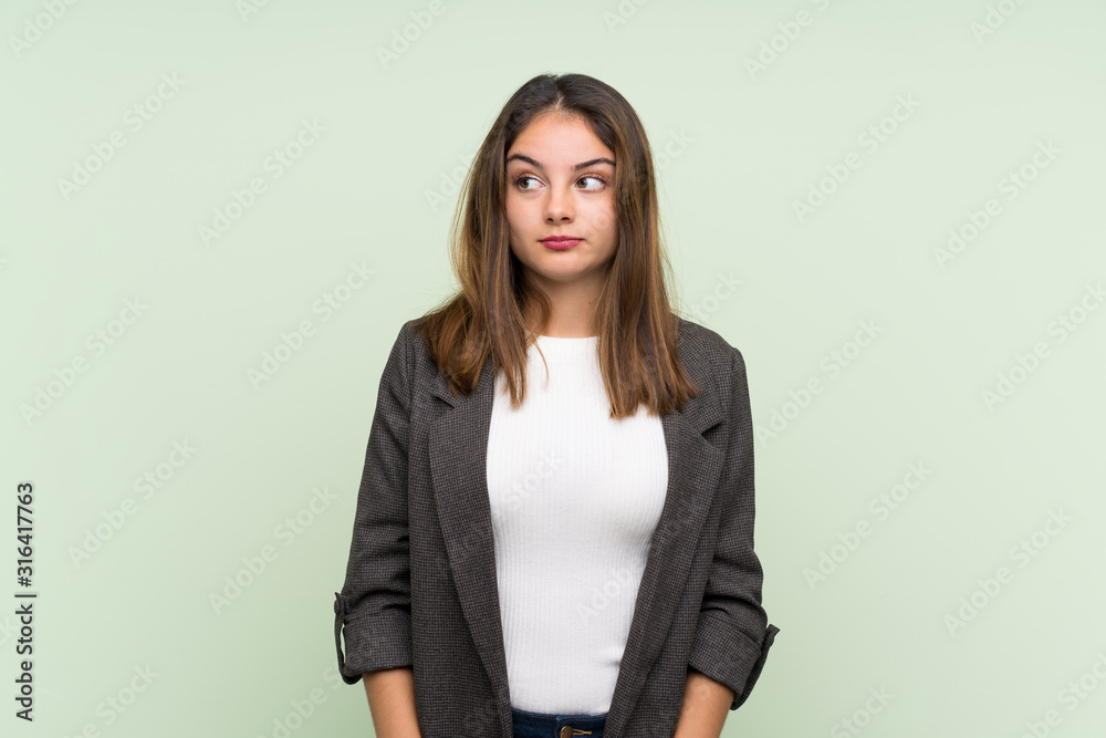 Young brunette girl with blazer over isolated green background making doubts gesture looking side Young brunette girl with blazer over isolated green background making doubts gesture looking side