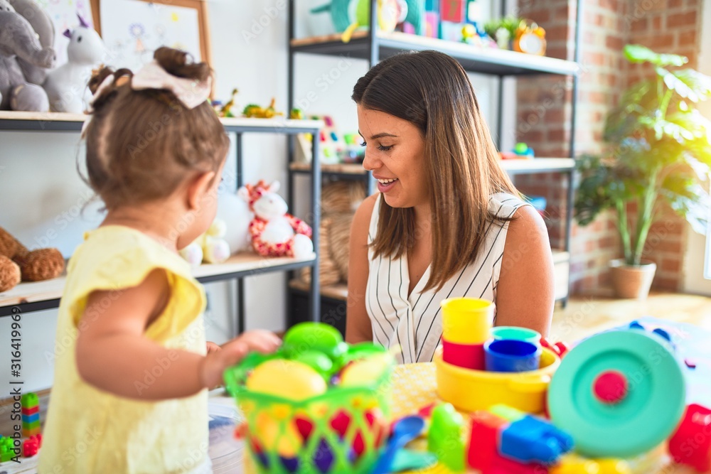 Young beautiful teacher and toddler playing with dishes, cutlery and cups toy on the table at kindergarten Young beautiful teacher and toddler playing with dishes, cutlery and cups toy on the table at kindergarten