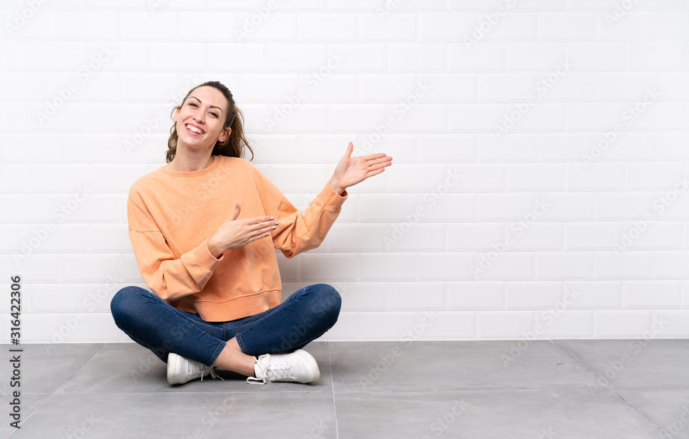 Young woman with curly hair sitting on the floor extending hands to the side for inviting to come Young woman with curly hair sitting on the floor extending hands to the side for inviting to come