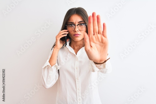 Young businesswoman wearing glasses talking on smartphone over isolated white background with open hand doing stop sign with serious and confident expression, defense gesture