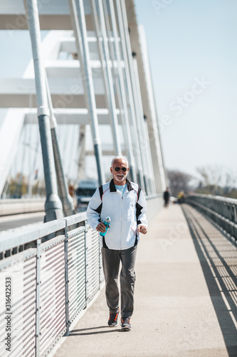 Fit senior man in good shape running and jogging alone on city bridge street. He is smiling and wears sunglasses.