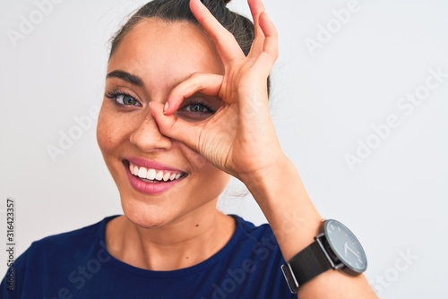 Young beautiful woman wearing blue casual t-shirt standing over isolated white background with happy face smiling doing ok sign with hand on eye looking through fingers