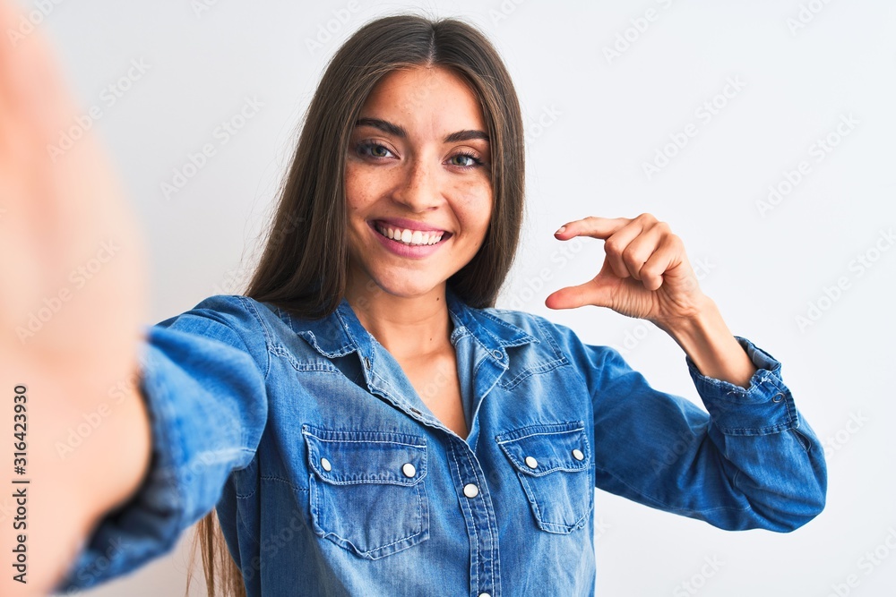Beautiful woman wearing denim shirt make selfie by camera over isolated white background smiling and confident gesturing with hand doing small size sign with fingers looking and the camera. Measure Beautiful woman wearing denim shirt make selfie by camera over isolated white background smiling and confident gesturing with hand doing small size sign with fingers looking and the camera. Measure