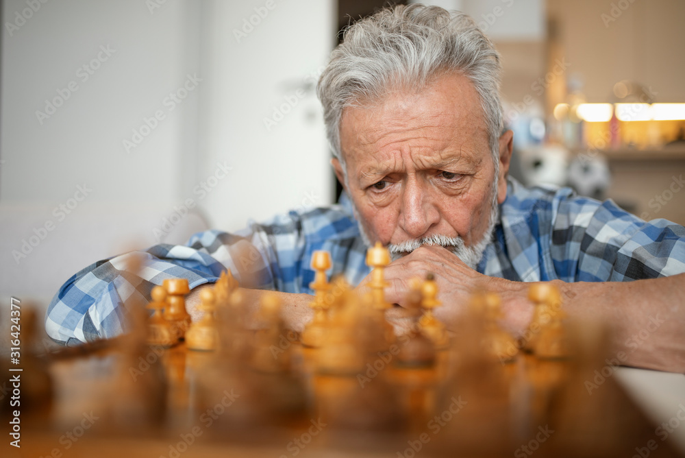 Chess stimulates his mind. Shot of an elderly man playing a game of chess by himself at home. Senior man contemplating chess move. Elderly man making a move with chess piece Chess stimulates his mind. Shot of an elderly man playing a game of chess by himself at home. Senior man contemplating chess move. Elderly man making a move with chess piece