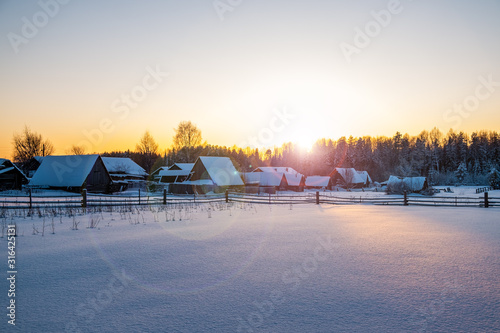 rural winter landscape. The sun sets behind the forest, creating highlights in the picture. Behind the fence are abandoned village houses the roofs are covered with snow