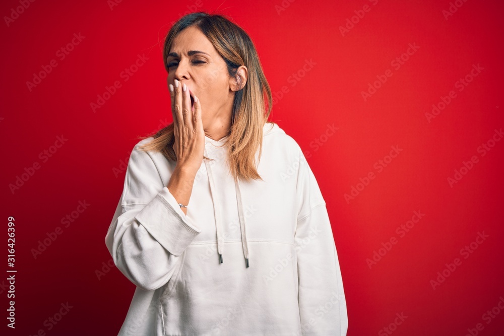 Middle age beautiful sportswoman wearing casual sweatshirt over isolated red background bored yawning tired covering mouth with hand. Restless and sleepiness. Middle age beautiful sportswoman wearing casual sweatshirt over isolated red background bored yawning tired covering mouth with hand. Restless and sleepiness.