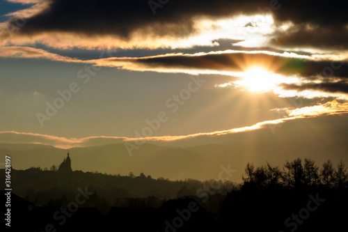 Majestic yellow sunset in the mountains landscape. Dramatic sky clouds, partly clear.sun hides in clouds,illuminating. mountains with forests on hills,a Church domes and roofs on horizon. evening time