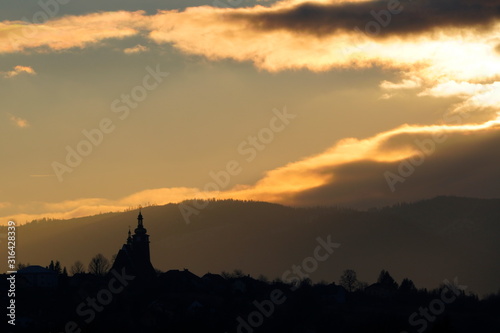 Majestic yellow sunset in the mountains landscape. Dramatic sky clouds, partly clear.sun hides in clouds,illuminating. mountains with forests on hills,a Church domes and roofs on horizon. evening time