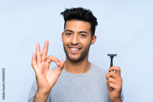 Young man shaving his beard over isolated blue background showing ok sign with fingers