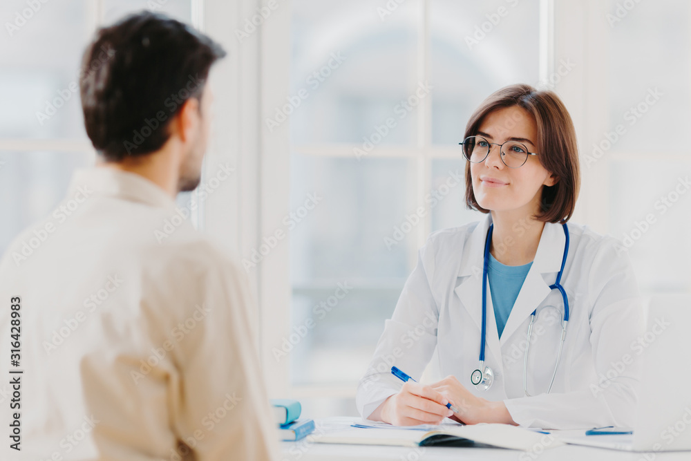 Heathcare, therapy session and assistance concept. Professional woman doctor speaks with male patient, gives consultancy and makes prescription, finds out symptoms of disease, provides help.