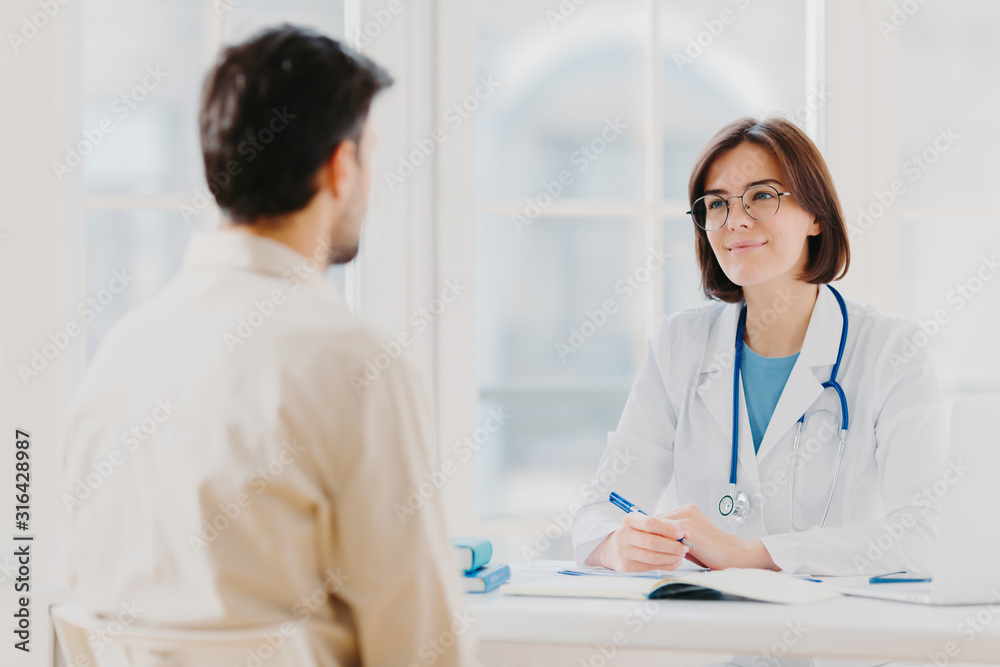 Doctor and patient discuss something, sit at table in clinic. Female cardiologist in eyewear gives medical consultation diagnostic, advice for man how to cure disease, pose in hospital room. Doctor and patient discuss something, sit at table in clinic. Female cardiologist in eyewear gives medical consultation diagnostic, advice for man how to cure disease, pose in hospital room.