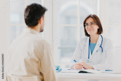 Doctor and patient discuss something, sit at table in clinic. Female cardiologist in eyewear gives medical consultation diagnostic, advice for man how to cure disease, pose in hospital room.