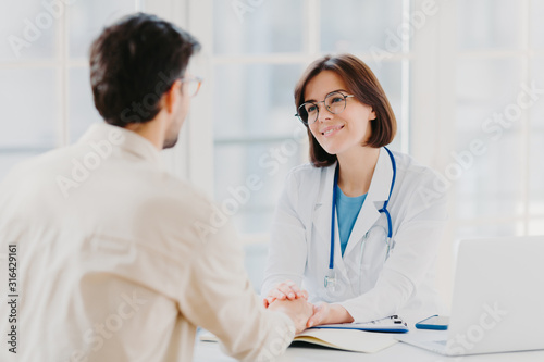 Confident female doctor holds hands of ill patient  persuades everything will be alright  dressed in white medical gown  gives advice  pose in hospial office. Consultation and diagnosis concept