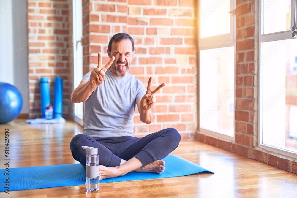 Middle age handsome sportman sitting on mat doing stretching yoga exercise at gym smiling with tongue out showing fingers of both hands doing victory sign. Number two.