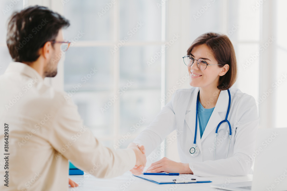 Friendly female doctor greets patient with handshake, pose in private clinic, sit opposite each other, have talk, agree to sign insurance contract, meet in hospital. Health care and medicine concept
