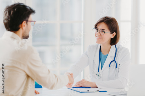 Friendly female doctor greets patient with handshake  pose in private clinic  sit opposite each other  have talk  agree to sign insurance contract  meet in hospital. Health care and medicine concept