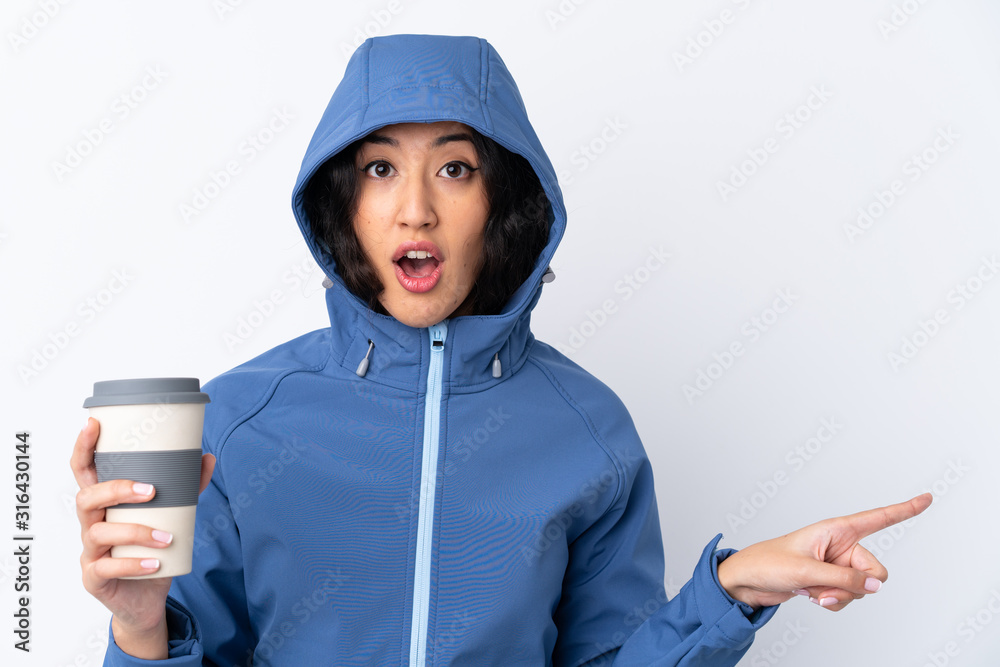 Mixed race woman wearing winter clothes with the hood on and holding a hot takeaway coffee over isolated white background surprised and pointing side Mixed race woman wearing winter clothes with the hood on and holding a hot takeaway coffee over isolated white background surprised and pointing side