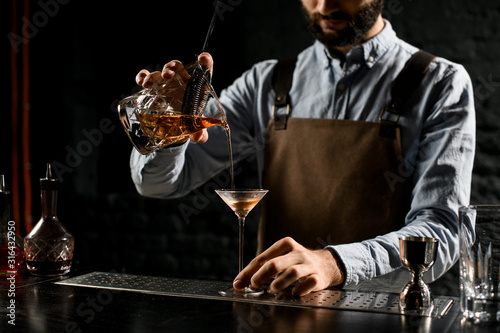 Professional male bartender pouring a brown alcoholic drink from the measuring cup with straner to a martini glass photo