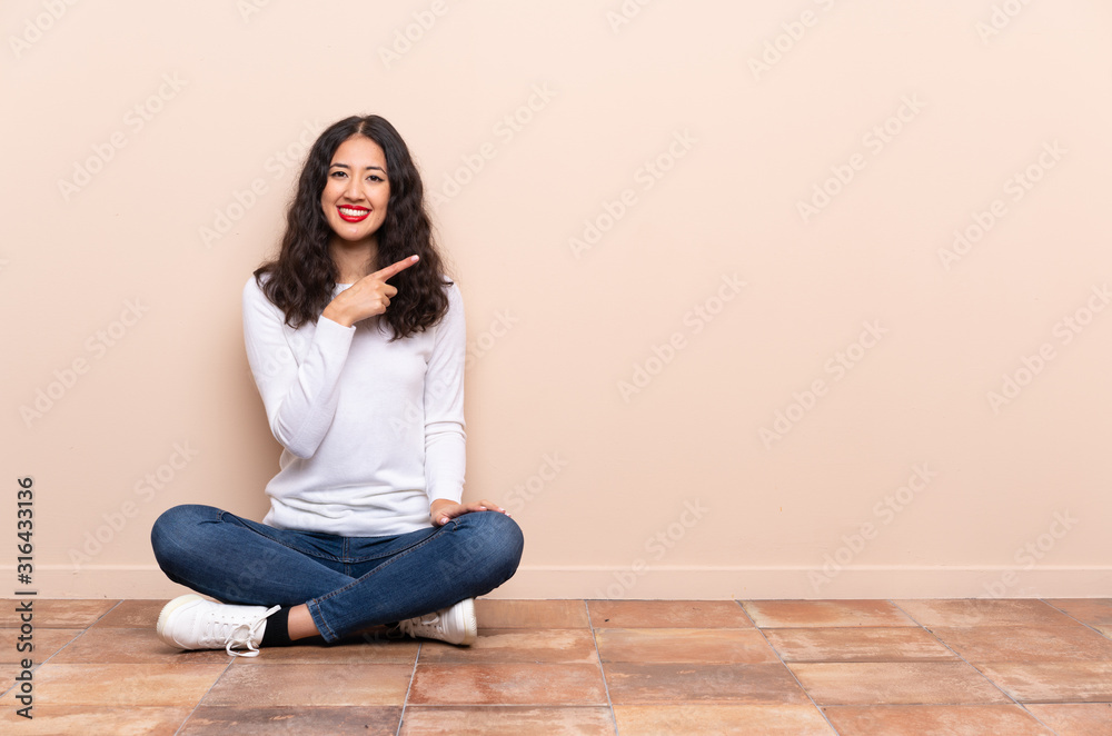 Young woman sitting on the floor pointing to the side to present a product - obrazy, fototapety, plakaty 