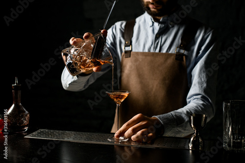 Bartender pouring a brown alcoholic drink from the measuring cup with straner to a martini glass