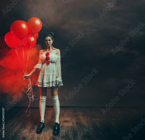 Obraz na płótnie portrait cute young woman with scary face clown grimm in white old dress stands in backdrop dark black gothic room holding red balloons