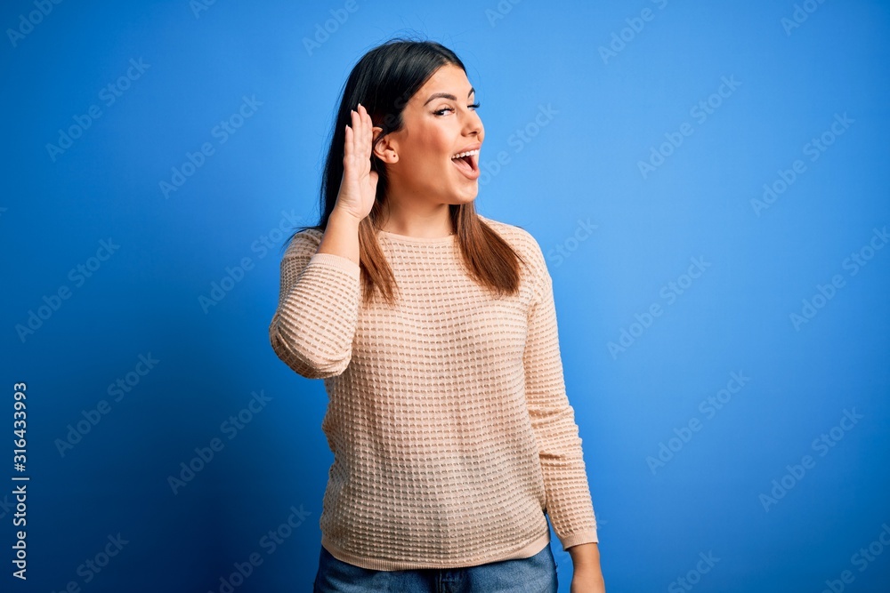 Young beautiful woman wearing casual sweater over blue background smiling with hand over ear listening an hearing to rumor or gossip. Deafness concept. Young beautiful woman wearing casual sweater over blue background smiling with hand over ear listening an hearing to rumor or gossip. Deafness concept.