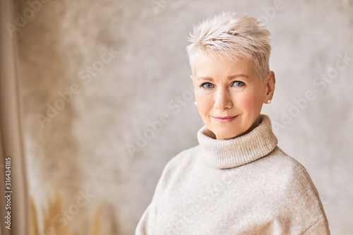фотографія Horizontal shot of blonde fifty year old European female with stylish pixie haircut and blue eyes smiling at camera, having confident look posing isolated indoors