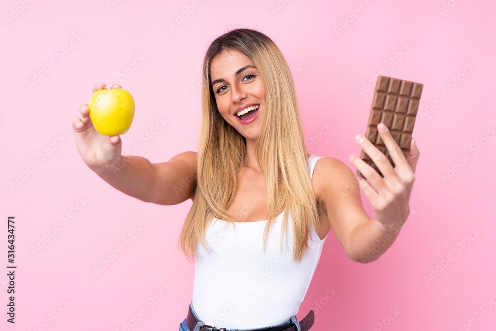 Obraz premium Young Uruguayan woman over isolated pink background taking a chocolate tablet in one hand and an apple in the other