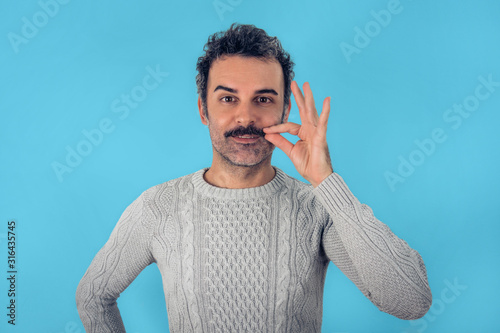 Funny portrait of brown, smiling, handsome man touching his mustache with gray sweater. Blue background. photo