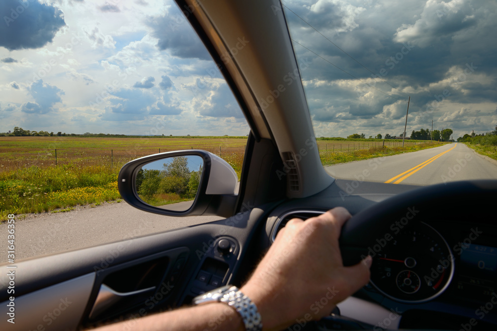 Man with hand on steering wheel of car driving down a country road with clouds Man with hand on steering wheel of car driving down a country road with clouds