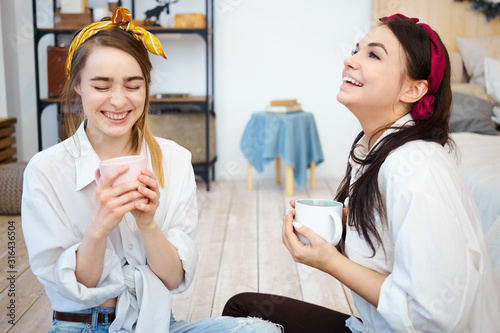 Pretty joyful girls having fun together indoors, sitting on floor with mugs of coffee, laughing, telling jokes and funny stories. Beautiful young women spending nice time, drinking tea at home