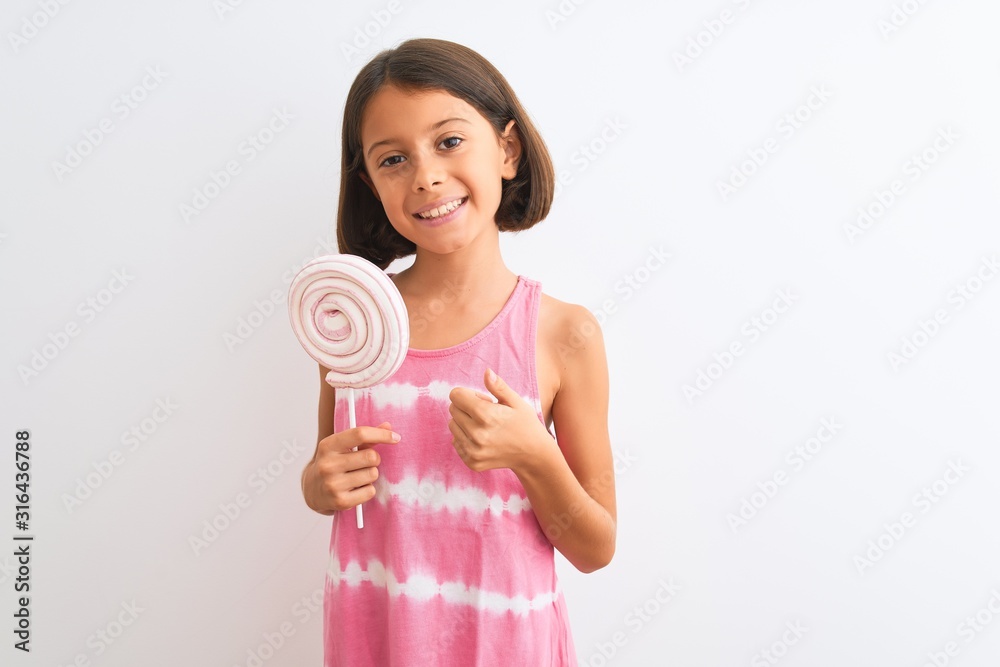 Beautiful child girl eating sweet lollipop standing over isolated white background happy with big smile doing ok sign, thumb up with fingers, excellent sign Beautiful child girl eating sweet lollipop standing over isolated white background happy with big smile doing ok sign, thumb up with fingers, excellent sign