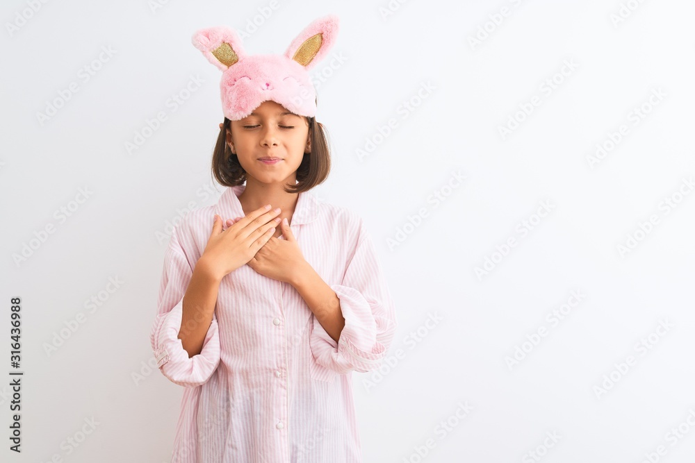 Beautiful child girl wearing sleep mask and pajama standing over isolated white background smiling with hands on chest with closed eyes and grateful gesture on face. Health concept.