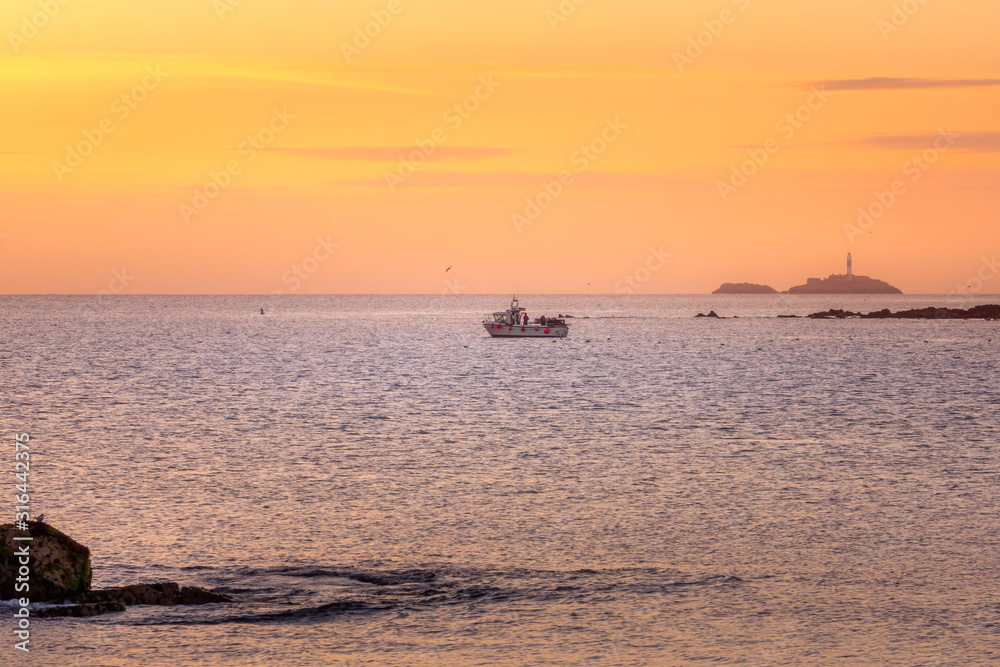 Fishing boat exiting port followed by flying and floating seagulls. Rockabill lighthouse on island in distance, golden hour sunrise, Skerries, Ireland