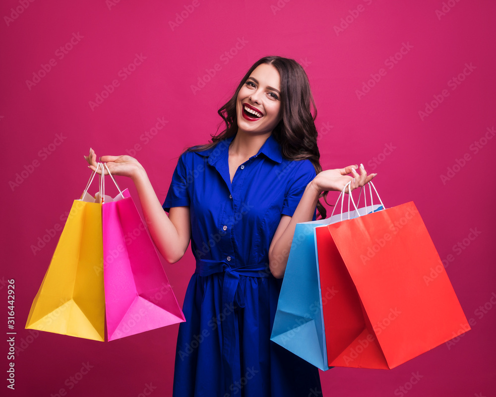 Brunette caucasian woman in blue dress on ponk background. She holds paper shopping bags in her hands and smiles happily. Emotional portrait with copy space