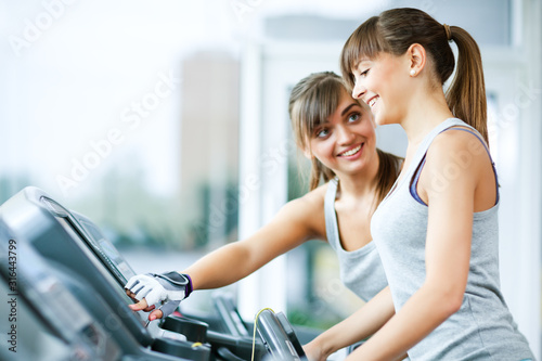 Young beautiful smiling woman in sportswear helping another girl with instructions for using treadmill in gym. Healthy active lifestyle, sport, bodycare concept