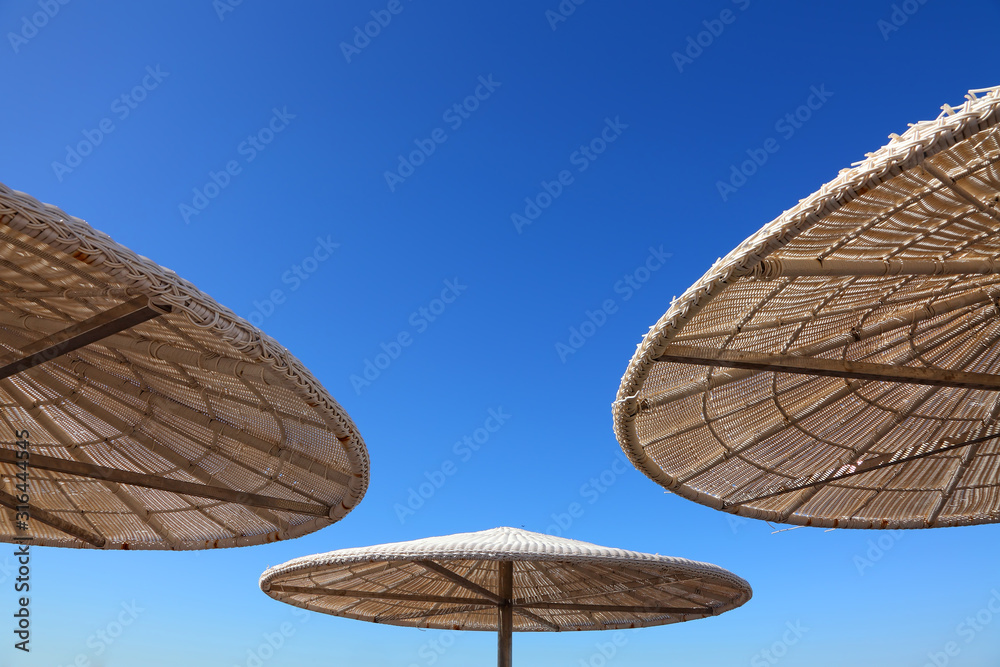 Fototapeta premium Tropical beach sun shades against bright blue sky on a sunny day. Up view. Sun umbrellas of reed and straw. Travel and vacation. Background with copy space