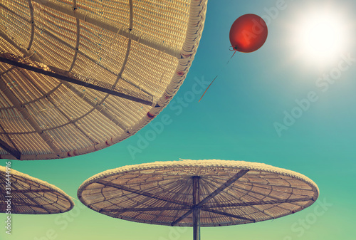 Tropical beach sun shades against blue sky. A red balloon flies to the sun shining glow. Up view. Sun umbrellas of reed and straw. Travel and vacation