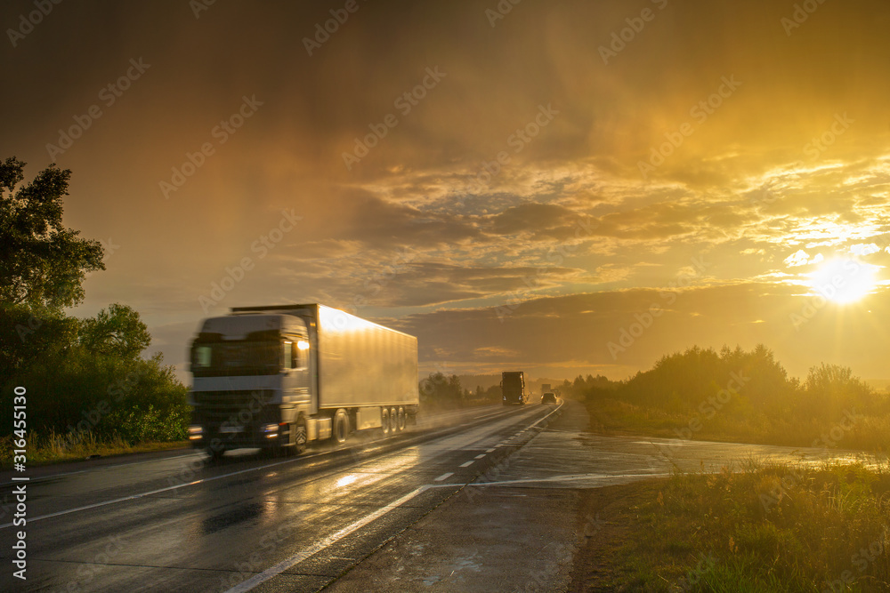 During an evening summer showers. A bright sun sunshine through thunderstorm clouds over the highway. Trucks and passenger cars go on the wet asphalt. Montage or graphic software was not used